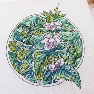 May include: A cross-stitch embroidery of a botanical design featuring green leaves and pink flowers within a circular frame. The artwork is on a white fabric, with a wooden frame partially visible.