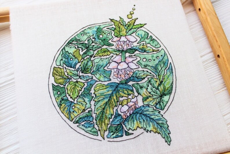 May include: A cross-stitch embroidery of a botanical design featuring green leaves and pink flowers within a circular frame. The artwork is on a white fabric, with a wooden frame partially visible.