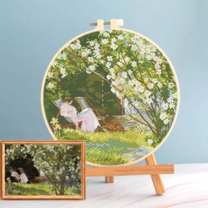 May include: A cross-stitch embroidery hoop featuring a pastoral scene of a person relaxing under a flowering tree. A smaller framed print of the same image is displayed in front. The scene is rendered in shades of green, white, and pink.