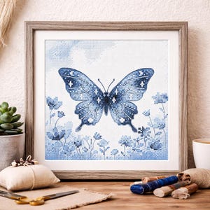 Può includere: Un'opera d'arte a punto croce incorniciata con una grande farfalla blu con dettagli di stelle e luna. La farfalla è circondata da fiori blu su sfondo bianco. L'opera è in una cornice di legno.