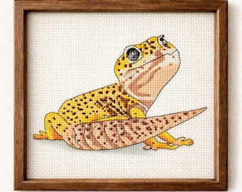 May include: A framed cross-stitch of a yellow and brown leopard gecko. The gecko has black spots and is set against a white background. The frame is made of dark brown wood. The gecko is looking up.