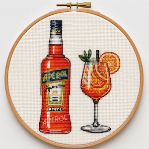 Könnte beinhalten: Eine Kreuzstichstickerei in einem runden Holzrahmen, die eine leuchtend orangefarbene Aperol-Flasche mit blau-weißem Etikett sowie ein Stielglas mit einem orangefarbenen Cocktail, garniert mit einer Orangenscheibe und grüner Minze, zeigt.