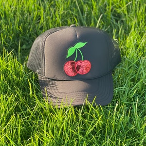 May include: A black mesh trucker hat with a red and green cherry patch. The cherry is made of sequins.