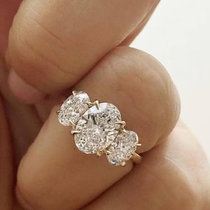 May include: A gold ring featuring three oval-cut diamonds. The center diamond is larger than the two side stones. The ring is set in a classic prong setting, showcasing the brilliance of the diamonds. The ring is held by a person.