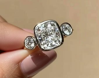 Puede incluir: Un anillo de oro con un gran diamante central de talla cojín cuadrado, flanqueado por dos diamantes redondos más pequeños. Los diamantes están engastados en una montura de platino u oro blanco, creando un diseño clásico y elegante.