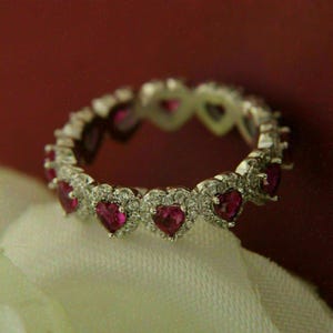 May include: A silver ring featuring heart-shaped ruby gemstones surrounded by small, clear accent stones. The ring is set against a red background and rests on a white fabric.