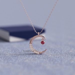 May include: A rose gold necklace featuring a crescent moon pendant. The moon is partially set with small clear stones along its curve and includes a single dangling round red stone. The text Kureightjewel is visible near the pendant base.