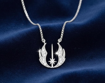 May include: A silver-colored pendant necklace featuring a stylized design with wings, a central sword, and a star. The pendant is encrusted with small, sparkling stones. The necklace chain is also silver and rests on a dark blue fabric.