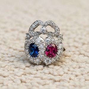 Puede incluir: Un anillo plateado con dos engastes en forma de corazón, uno con una piedra preciosa azul oscuro y otro con una piedra preciosa roja vibrante, rodeados de pequeñas piedras transparentes. El anillo tiene un diseño en forma de lazo en la parte superior, también adornado con piedras transparentes.