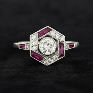 May include: A platinum ring featuring a central round brilliant-cut diamond surrounded by a hexagonal frame. The frame is accented with alternating baguette-cut red rubies and small round diamonds, set in a milgrain-edged metal setting.