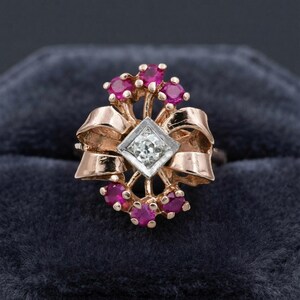May include: A rose gold ring featuring a central square-set diamond surrounded by a ribbon-like metal design. The piece is accented with six round-cut vibrant pink rubies arranged in clusters at the top and bottom of the central diamond setting.