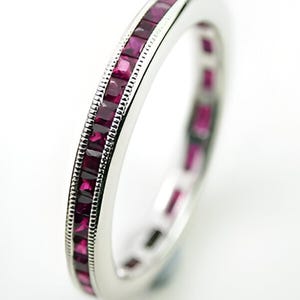 May include: A silver ring with a continuous band of square-cut, deep red gemstones. The ring has a detailed, textured edge, and the gemstones are set closely together, creating a vibrant, eye-catching design. The ring is set against a bright white background.