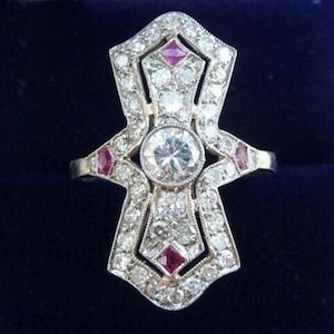 May include: An Art Deco style ring with a central round diamond and smaller diamonds and rubies. The ring has a geometric design with a silver band and is set against a dark blue background.