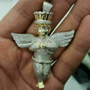 May include: A gold and silver pendant in the shape of a cherub with a crown. The cherub has outstretched wings and is covered in small, sparkling stones. The pendant is held in a hand.