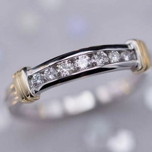 Puede incluir: Un anillo de plata y oro con siete piedras preciosas redondas y transparentes engastadas en un canal. El anillo tiene un diseño curvo con detalles dorados en cada extremo. Las piedras preciosas brillan.