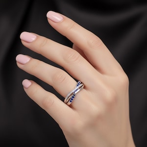 May include: A silver-toned crossover ring featuring two intertwined bands. One band is encrusted with small, clear white stones, while the other band is set with small, deep blue stones. The ring has a polished metallic finish and a split-shank design.