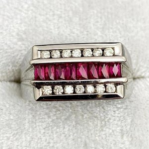 May include: A silver-toned ring featuring a central horizontal row of seven rectangular-cut deep red rubies flanked by two parallel rows of six small round-cut clear white diamonds, all set within a polished metal band with a channel setting.