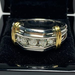 May include: A two-tone ring featuring a polished silver-colored band with a central channel set with five round clear stones. The ring is accented by two vertical gold-colored bars on either side of the stone setting, creating a structured design.