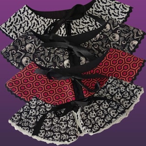 Puede incluir: Cuatro cuellos decorativos desmontables con patrones únicos. Los cuellos presentan lazos de cinta negra y ribete de encaje. Los patrones incluyen murciélagos, calaveras, huesos y un diseño geométrico rojo y negro. Los cuellos están superpuestos sobre un fondo púrpura.
