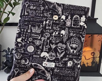 May include: A black book cover featuring white occult symbols, keys, and script. It has a button closure and a label reading "Rosemary Couture". The design includes crowns, eyes, and other mystical elements, with a button closure.
