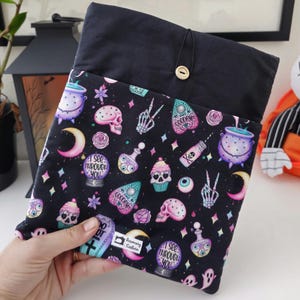 May include: Black fabric book sleeve with a button closure. The patterned fabric features skulls, potion bottles, moons, and the text "I SEE THROUGH YOU" in a spooky theme. The top is solid black.