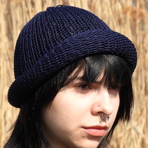 May include: A navy blue knitted beanie hat with a short brim. The hat is made of a textured, woven material. The person in the image is wearing a black turtleneck.