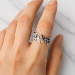 Może przedstawiać: Srebrny pierścionek z motywem skrzydła smoka. Pierścionek posiada dwa stylizowane skrzydła, które oplatają palec. Skrzydła mają szczegółowe tekstury i ciemne, utlenione wykończenie, tworząc gotycką lub fantastyczną estetykę.