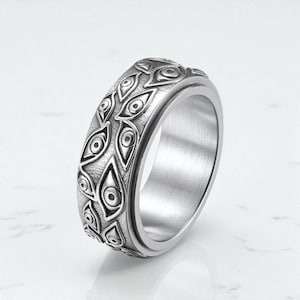 May include: A silver ring featuring a repeating pattern of eye motifs. The ring has a polished finish and a spinning band. The eye design is detailed and wraps around the entire ring, creating a distinctive piece of jewellery.