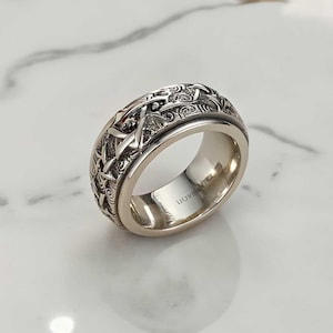 Könnte beinhalten: Ein silberner Ring mit einem aufwendigen, strukturierten Design. Der Ring hat ein detailliertes Muster aus ineinander verschlungenen Formen und Wirbeln um das Band. Das Innere des Rings ist glatt und poliert, mit dem Text "LIOVRISKI" sichtbar.