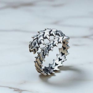 May include: A silver ring with a detailed, articulated design resembling a spine or segmented creature. The ring is shown on a white marble surface, with the ring's intricate details catching the light.