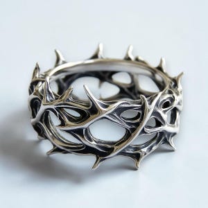 May include: A silver-toned ring featuring a continuous band design of interconnected, sharp, thorn-like protrusions. The metal has a polished finish with darkened, recessed areas that emphasize the intricate, organic, and jagged structure of the band.
