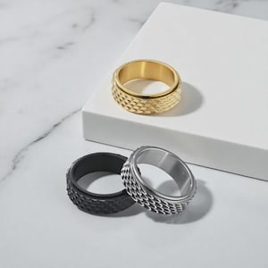 Peut inclure: Trois bagues en métal texturé, dorées, noires et argentées. La bague dorée est posée sur une surface blanche, tandis que les bagues noire et argentée sont côte à côte en dessous. Les bagues ont un motif répétitif.