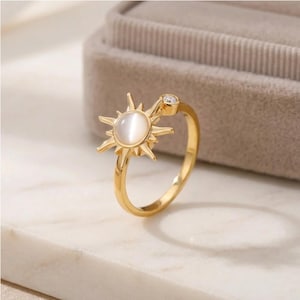 Puede incluir: Un anillo de tono dorado con un motivo central de sol que presenta una piedra cabujón redonda de color blanco lechoso en el centro. El diseño solar incluye rayos radiantes y una pequeña piedra redonda transparente engastada en el aro.
