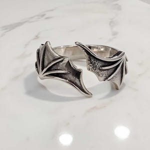 May include: A silver-colored ring with a bat wing design. The ring features two detailed bat wings on either side, with intricate vein detailing. The ring is set against a white marble surface.