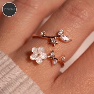 Pode incluir: Um anel aberto em tom de ouro rosa com um design de ramo, pequenas pedras transparentes e um elemento floral branco. A banda tem a forma de um raminho com folhas e pétalas, acentuada por pedras brilhantes. O texto LYNGIA é visível no canto.