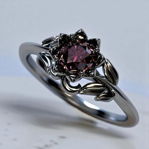 Op de afbeelding: Zilverkleurige ring met een bloemvormige zetting met een dieprode edelsteen. De ring heeft een bloemmotief met bladeren en een fijne band, wat een natuurlijke uitstraling geeft. Een sieraad.