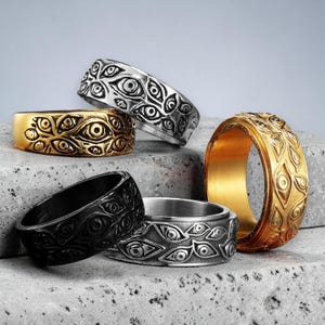 Op de afbeelding: Een verzameling van vier metalen ringen met een reliëfpatroon van meerdere ogen over het gehele oppervlak. De set bevat één zilverkleurige ring, één zwarte ring en twee goudkleurige ringen, die allemaal een consistent oogmotief vertonen.