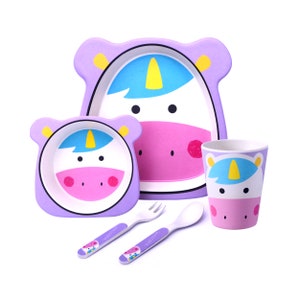 May include: A children's unicorn-themed dinner set. The set includes a plate, bowl, cup, fork, and spoon. The items are primarily lavender, white, pink, and yellow. The plate and bowl are shaped like a unicorn head. The fork and spoon have the word "unicorn" printed on them.