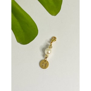 Op de afbeelding: Gouden bedel met een enkele witte parel en een gouden schijf met de letter 'H' erop gegraveerd.