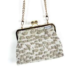 Peut inclure: Un petit sac à main à motifs avec une bandoulière en chaîne dorée. Le sac est doté d'une fermeture à fermoir et d'un tissu texturé blanc cassé avec un motif répété de formes grises. La marque "kadomaro" est visible sur une petite étiquette.