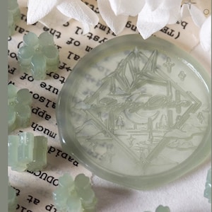 May include: Two round, translucent green wax seals with raised designs. One seal features a crescent moon and a stylised landscape, the other features a mountain range and a stylised building. The seals are surrounded by small, white and green beads.