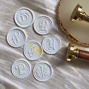 May include: A set of eight white wax seals with different letter initials, including D, R, S, F, H, T, P, and I. The seals are arranged on a white fabric surface. Two gold-toned wax seal stamps are also shown.