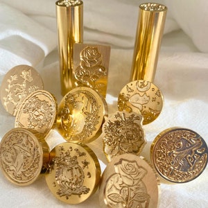 May include: A collection of ten gold-toned metal wax seals with various designs, including a skull, a rose, a crescent moon, a cat, and a cauldron. The seals are arranged on a white fabric surface. Two gold-toned metal handles are also included.