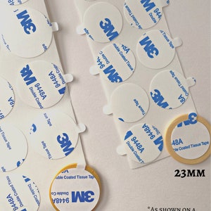 May include: Close-up of two different sizes of round, white, adhesive 3M double-coated tissue tape. The larger tape is 28mm in diameter and the smaller tape is 23mm in diameter. The text "AS SHOWN ON A SEAL MADE WITH A 25MM STAMP" is printed below the tape.