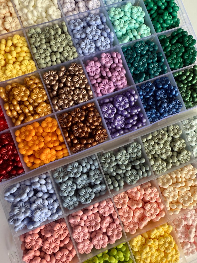 May include: A close-up view of a storage box filled with various colors of small flower-shaped beads. The beads are arranged in rows and columns, creating a colorful and organized display.