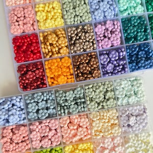 May include: A clear plastic storage box filled with small flower-shaped beads in various colors, including red, orange, yellow, green, blue, purple, and white.