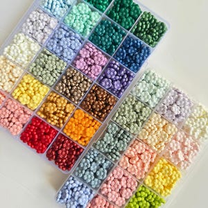 May include: A clear plastic storage box filled with small, colorful flower-shaped beads. The beads are arranged in rows by color, with shades of green, blue, purple, gold, orange, red, and pink.