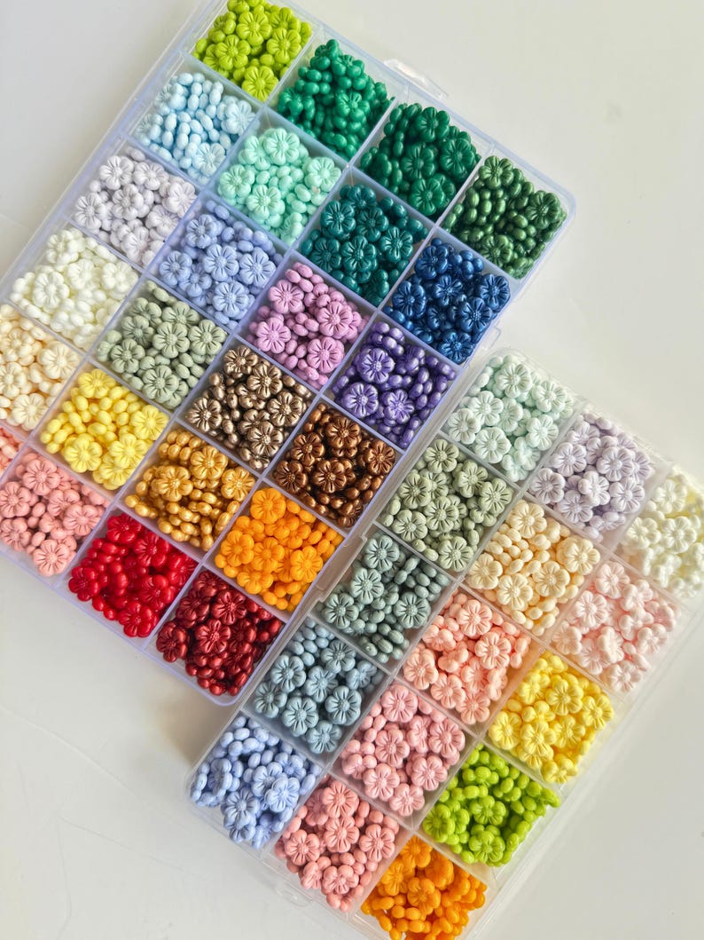 May include: A clear plastic storage box filled with small, colorful flower-shaped beads. The beads are arranged in rows by color, with shades of green, blue, purple, gold, orange, red, and pink.