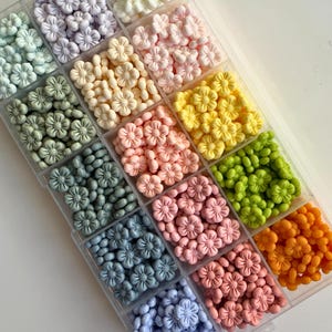 May include: A clear plastic storage box filled with small, pastel-colored flower-shaped beads. The beads are arranged in rows, with each row containing a different color. The colors include white, light purple, light yellow, light pink, light green, light blue, and light orange.