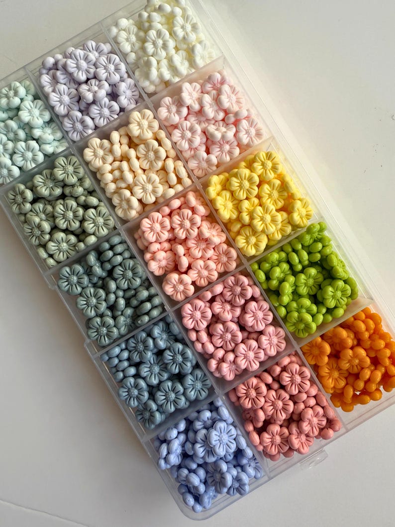 May include: A clear plastic storage box filled with small, pastel-colored flower-shaped beads. The beads are arranged in rows, with each row containing a different color. The colors include white, light purple, light yellow, light pink, light green, light blue, and light orange.